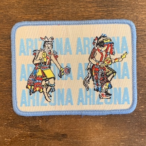 Peut inclure: Un écusson en tissu blanc avec une bordure bleue représentant deux danseurs amérindiens vêtus de vêtements colorés. L'écusson porte le texte "Arizona" répété plusieurs fois en bleu.