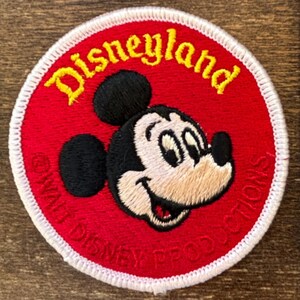 May include: Embroidered patch featuring Mickey Mouse with the word "Disneyland" in yellow script. The patch has a red background with a white border and the text "Walt Disney Productions".