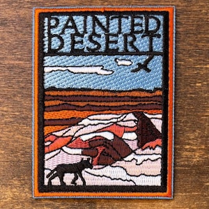 Puede incluir: Parche bordado con un diseño de paisaje desértico y la frase "PAINTED DESERT". El parche tiene un borde negro con un ribete naranja. El diseño incluye un cielo azul, nubes, una escena desértica y una silueta negra de coyote.