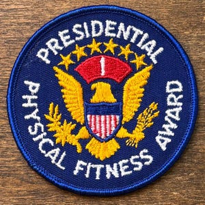 May include: A circular blue patch with white text that reads "Presidential Physical Fitness Award." The patch features a gold eagle with a red and white shield, a red banner, and gold stars.