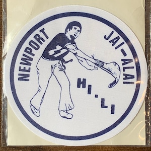 Peut inclure: Un patch circulaire blanc avec une bordure bleue et une illustration bleue d'une personne jouant au jai alai. Le texte "NEWPORT JAI-ALAI HI-LI" est écrit autour de la bordure.