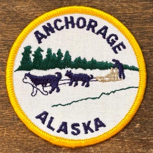 Pode incluir: Patch bordado circular com fundo branco, com o texto "ANCHORAGE ALASKA" em azul marinho. O patch tem uma borda amarela e retrata uma equipe de trenó puxado por cães, árvores e uma pessoa.
