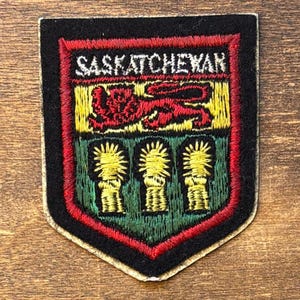 May include: A shield-shaped embroidered patch featuring the word SASKATCHEWAN in white thread at the top. The design includes a red lion on a yellow background above three yellow wheat sheaves on a green background, all bordered by red and black.