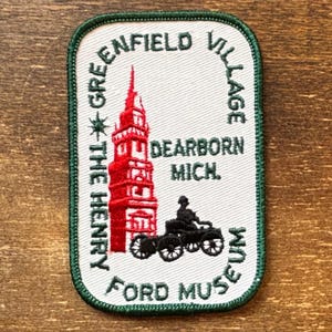 May include: A rectangular embroidered patch with a dark green border on a white background. It features a red tower, a black vintage car, and black text reading THE HENRY FORD MUSEUM, GREENFIELD VILLAGE, and DEARBORN MICH. The design is centred.