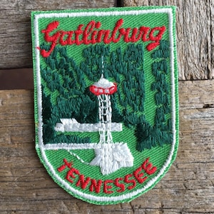 May include: Embroidered patch featuring the text "Gatlinburg Tennessee" in red script. The patch has a green background with a white and red tower design, surrounded by green trees. The patch is bordered with white stitching.