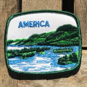 Op de afbeelding: Een witte geborduurde patch met een groene rand. De patch toont een landschap met een rivier, groene heuvels en het woord "AMERICA" in blauwe borduurwerk.