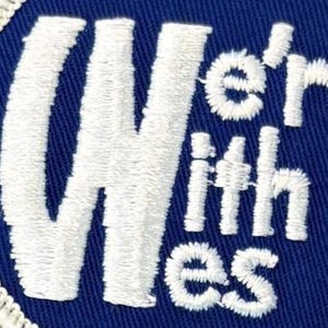 Puede incluir: Un parche bordado circular que presenta un fondo de tela azul profundo con el texto bordado en blanco We're With Wes. El bordado tiene una textura en relieve y est&aacute; acabado con un borde cosido en blanco alrededor del borde exterior circular.