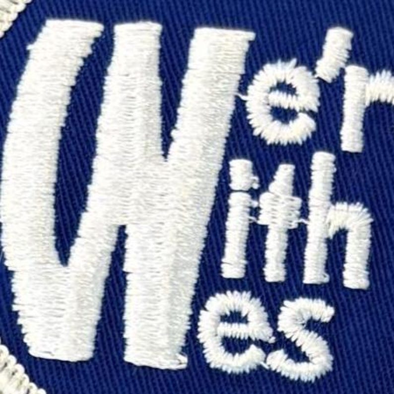 Puede incluir: Un parche bordado circular que presenta un fondo de tela azul profundo con el texto bordado en blanco We're With Wes. El bordado tiene una textura en relieve y est&aacute; acabado con un borde cosido en blanco alrededor del borde exterior circular.