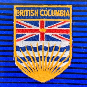 Könnte beinhalten: Ein blauer und gelber gestickter Aufnäher mit dem Text "BRITISH COLUMBIA" und einem stilisierten Bild der Flagge von British Columbia. Die Flagge zeigt im oberen linken Eck eine Union Jack und im unteren Teil ein Sonnenstrahlen-Design.