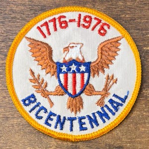 Peut inclure: Écusson brodé de forme circulaire avec un motif patriotique. Il présente un aigle, un bouclier à rayures rouges et blanches, et trois étoiles. Les inscriptions "1776-1976" et "BICENTENNIAL" sont en rouge et bleu sur fond blanc, avec une bordure jaune.