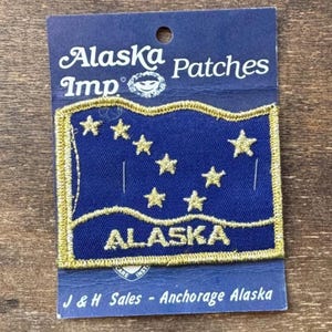 以下が含まれることがあります： 金色の縁取りと7つの金色の星が付いた青いアラスカのパッチで、金色の文字で「ALASKA」と書かれています。パッチは、「Alaska Patches」と「J＆H Sales-Anchorage Alaska」という言葉が書かれた青いカードにあります。