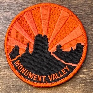 May include: A circular embroidered patch featuring a black silhouette of the Monument Valley landscape against a bright orange sunburst background. The text MONUMENT VALLEY is stitched in orange along the bottom edge of the black terrain.