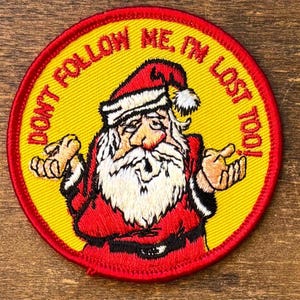 May include: Embroidered patch featuring Santa Claus with a confused expression. The patch is circular, with a yellow center and red border. Text around the edge reads "DON'T FOLLOW ME, I'M LOST TOO!"