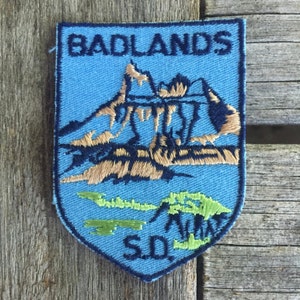 Könnte beinhalten: Blauer und brauner gestickter Aufnäher mit den Worten "Badlands SD" und einer Darstellung einer Bergkette.