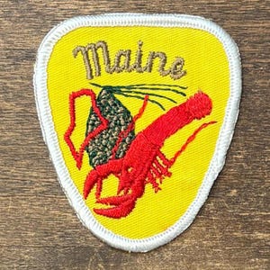 May include: A shield-shaped embroidered patch with a yellow background and a white border. It features the word Maine in brown script, a green pinecone with needles, a red lobster, and a red outline of the state of Maine on the yellow fabric.