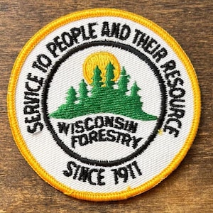 Op de afbeelding: Geborduurde patch met een witte kern, met een groen bos silhouet onder een gele zon. De patch heeft een zwarte rand met de tekst "SERVICE TO PEOPLE AND THEIR RESOURCE" en "WISCONSIN FORESTRY SINCE 1911". De buitenste rand is geel.