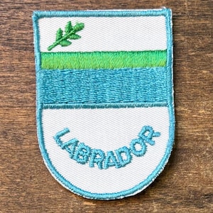 May include: Embroidered patch with a white background, featuring a green sprig, a green stripe, a blue stripe, and the word "LABRADOR" in blue. The patch has a blue border and is on a wooden surface.