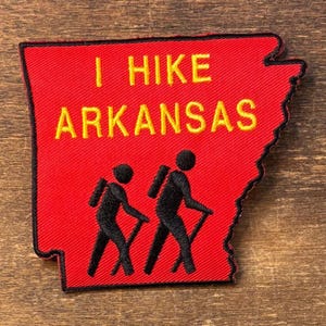 May include: A red embroidered patch shaped like the state of Arkansas with a black border. The text I HIKE ARKANSAS is stitched in yellow. Below the text are two black silhouettes of hikers carrying backpacks and using trekking poles.