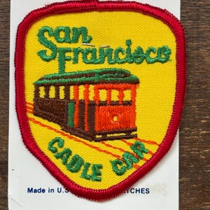 Op de afbeelding: Geborduurde patch met een San Francisco kabeltram ontwerp. De patch heeft een gele achtergrond en een rode rand. De tekst "San Francisco Cable Car" is groen. De kabeltram is oranje en bruin. Gemaakt in de VS.