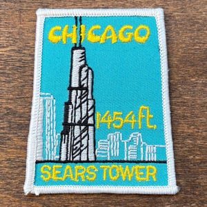 Puede incluir: Parche bordado con el horizonte de Chicago, incluyendo la Torre Sears, con la altura marcada como "1454 ft." en amarillo. El parche tiene un fondo turquesa con detalles blancos y negros, y el texto "CHICAGO" y "SEARS TOWER" en amarillo.