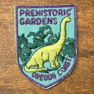May include: A shield-shaped embroidered patch with a light blue background and a purple border. It features a yellow long-necked dinosaur standing in front of dark green foliage. The text reads PREHISTORIC GARDENS at the top and OREGON COAST at the bottom.