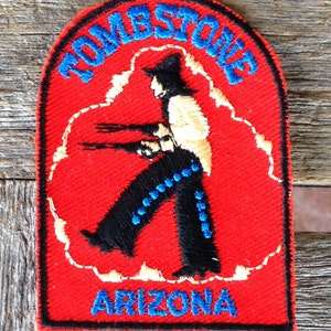 Puede incluir: Parche bordado con fondo rojo, borde negro y las palabras "TOMBSTONE ARIZONA" en azul. El parche presenta una figura con pantalones negros con detalles azules, camisa beige y sombrero negro, sosteniendo dos pistolas.