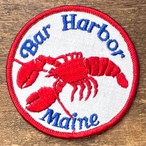 May include: A circular embroidered patch with a white background and a red border. The patch features a bright red lobster in the center. The blue text "Bar Harbor" is arched across the top, and the blue text "Maine" is at the bottom.