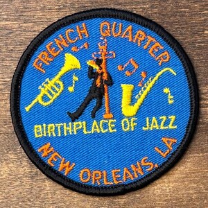 May include: A circular embroidered patch with a blue background and black border. It features a yellow trumpet, a yellow saxophone, and a black silhouette of a person playing an instrument. Text reads: FRENCH QUARTER BIRTHPLACE OF JAZZ NEW ORLEANS, LA.