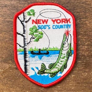 Puede incluir: Parche bordado con borde rojo que representa una escena de pesca. El diseño incluye un pez verde y blanco, un lago azul, un árbol y el texto "NEW YORK GOD'S COUNTRY" en rojo y verde.