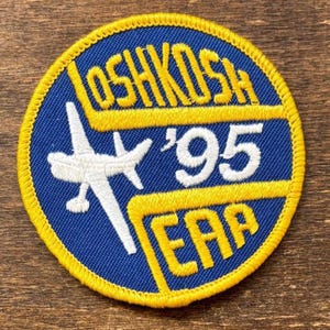 May include: A circular patch with a blue background and yellow trim. The patch features the text "OSHKOSH '95 EAA" in yellow, along with a white airplane graphic. The patch is likely a souvenir or collectible item.