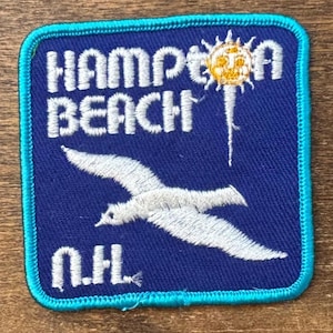May include: A square embroidered patch with a blue background and a bright turquoise border. It features white text reading HAMPTON BEACH, a yellow sun icon, a white seagull in flight, and the white text N.H. at the bottom left corner.