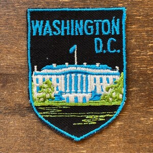 May include: Embroidered patch with the text "WASHINGTON D.C." in turquoise above a White House illustration. The patch has a black background with a turquoise border and green and white details. Ideal for jackets or bags.