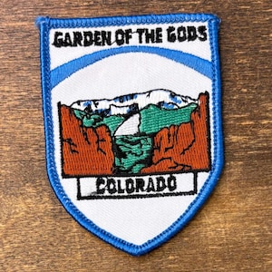 Op de afbeelding: Een schildvormige geborduurde patch met een blauwe rand. De witte achtergrond toont een landschap van rode rotsformaties, groene heuvels en besneeuwde bergen. De tekst GARDEN OF THE GODS staat bovenaan en COLORADO onderaan.