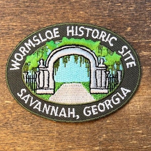May include: An embroidered patch featuring the Wormsloe Historic Site in Savannah, Georgia. The oval patch has a dark green border with white text. The image depicts a white archway, a path, and green foliage.