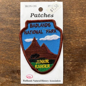 May include: A brown and blue embroidered patch with the text "Badlands National Park" and "Junior Ranger". The patch is on a white card with the text "Iron On Sew Patches $1.50" and "Badlands Natural History Association".