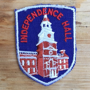 Peut inclure: Écusson brodé représentant l'Independence Hall de Philadelphie. L'écusson a un fond bleu avec des coutures blanches. Le bâtiment est rouge et blanc, avec les mots "INDEPENDENCE HALL" en rouge.