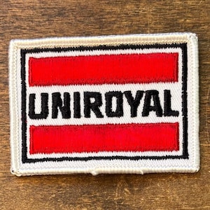Op de afbeelding: Geborduurde patch met het woord "UNIROYAL" in zwarte blokletters. De patch heeft een witte rand met een zwarte omtrek, die een rood rechthoekig ontwerp omlijnt. De patch is rechthoekig van vorm.
