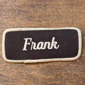 Peut inclure: Un écusson rectangulaire marron foncé avec le nom "Frank" brodé en blanc. Le patch a une bordure cousue beige. Le fond est une surface en bois.