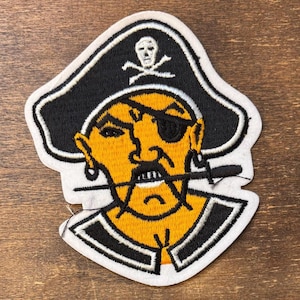 May include: An embroidered patch featuring a yellow pirate face wearing a black tricorn hat with a white skull and crossbones emblem. The pirate has a black eye patch, a stern expression, and a black sword held horizontally between the teeth.