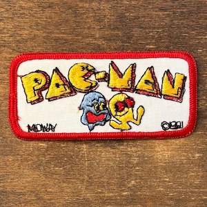 Könnte beinhalten: Ein rechteckiger, bestickter Aufnäher mit weißem Hintergrund und rotem Rand. Er zeigt den gelben Pac-Man-Schriftzug, einen blauen Geist und eine gelbe Pac-Man-Figur. Der Text Midway und ©1981 ist in schwarzem Garn eingestickt.