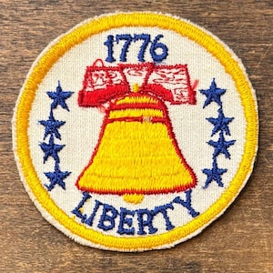 Peut inclure: Écusson brodé circulaire avec un bord jaune, représentant la Cloche de la Liberté jaune avec des détails rouges. Le texte "1776" est en bleu au-dessus de la cloche, et "LIBERTY" en dessous. Des étoiles bleues entourent le motif central.