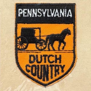 May include: A shield-shaped embroidered patch with a black border. The top black section features the white text PENNSYLVANIA. The orange lower section displays a black silhouette of a horse-drawn carriage and the black text DUTCH COUNTRY.