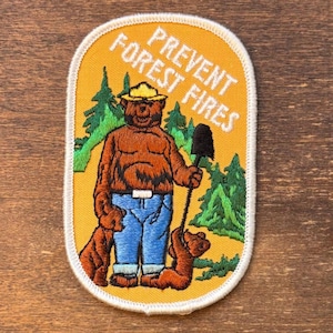 May include: Embroidered patch featuring Smokey Bear in a yellow hat and blue jeans, holding a shovel. The patch has the text "PREVENT FOREST FIRES" in white letters on a yellow background, with a white border.