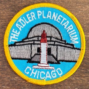 Puede incluir: Parche bordado circular con el Adler Planetarium de Chicago. El parche tiene un fondo azul claro y un borde amarillo. El edificio del planetario es gris con una torre roja y blanca. El texto dice "THE ADLER PLANETARIUM" y "CHICAGO".