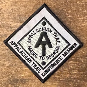 May include: A diamond-shaped embroidered patch with a white background and black border. It features the black Appalachian Trail logo in the center, with the text Appalachian Trail, Maine to Georgia, and Appalachian Trail Conference Member.