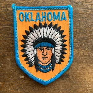 May include: An orange and blue embroidered patch with the word "OKLAHOMA" in blue lettering. The patch features a Native American man wearing a headdress with white feathers.
