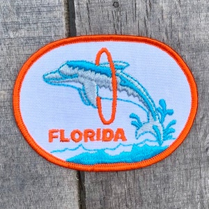 May include: An orange oval embroidered patch with a white background. The patch features a blue and gray dolphin jumping through a red hoop. The dolphin is surrounded by blue waves and the word "FLORIDA" is embroidered in orange below the dolphin.
