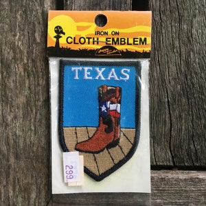 May include: An iron-on cloth emblem in original packaging featuring a shield-shaped patch with a blue background, the word TEXAS in white, and a brown cowboy boot with a Texas flag design standing on a tan wooden floor surface.
