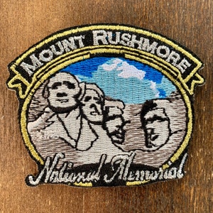 Könnte beinhalten: Gestickter Aufnäher mit dem Mount Rushmore National Memorial. Der Aufnäher hat einen schwarzen Rand mit den Worten "Mount Rushmore" und "National Memorial" in weißer Schrift. Das Bild zeigt die vier Präsidenten vor blauem Himmel mit weißen Wolken.