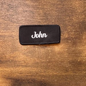 以下が含まれることがあります： 黒い長方形のパッチで、名前「John」が白い筆記体で刺繍されています。このパッチは、おそらく衣類用で、木製の背景に表示されています。